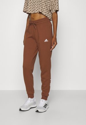 Person trägt braune Adidas-Sweathosen, weiße Adidas-Sneakers und ein bauchfreies Top mit geometrischem Muster.