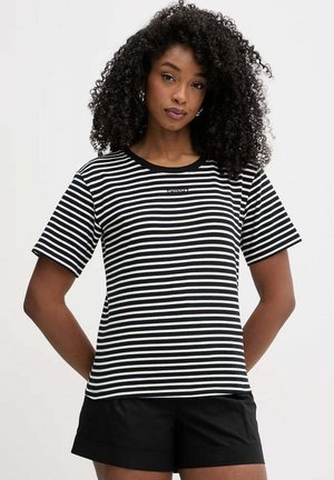 Donna con capelli ricci che indossa una maglietta a righe orizzontali bianca e nera e pantaloncini neri, in piedi con le mani dietro la schiena.