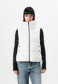 Vrouw in een witte puffer vest met hoge kraag over een zwart-wit gestreept lange mouwen top en lichtblauwe spijkerbroek, staand tegen een witte achtergrond.