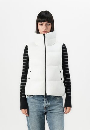 Vrouw in een witte puffer vest met hoge kraag over een zwart-wit gestreept lange mouwen top en lichtblauwe spijkerbroek, staand tegen een witte achtergrond.