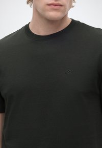 Jeune personne portant un t-shirt uni vert foncé à col rond avec un petit logo brodé sur le côté gauche de la poitrine.