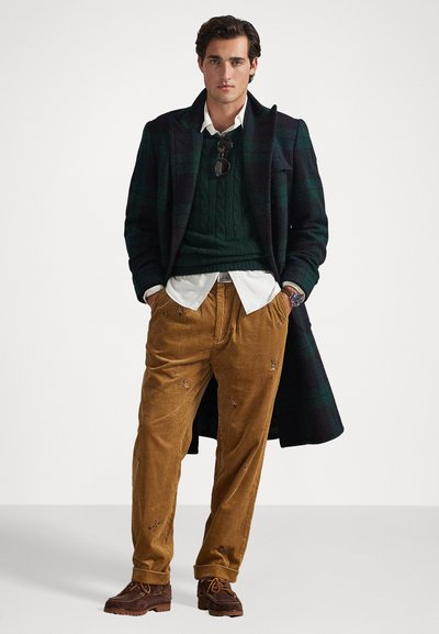 Geruite wollen overjas, donkergroene kabelgebreide trui, witte bloes met kraag, bruine corduroy broek met bloem borduursel, bruine veterschoenen.