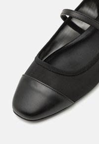 Black ballet flat featuring a textured fabric upper and a glossy leather toe cap. Includes a slim, decorative strap across the top.