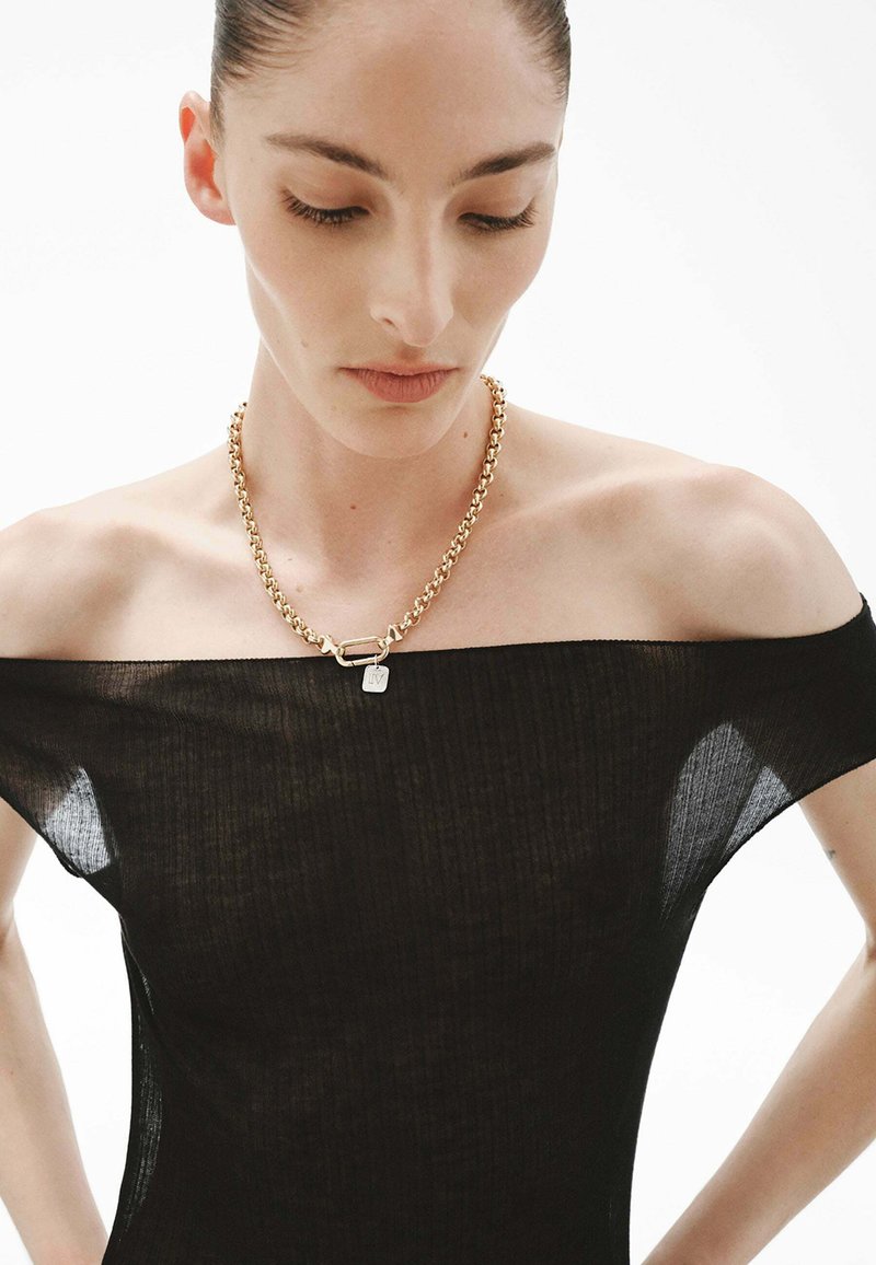 Collar de cadena de oro con un colgante cuadrado, que presenta eslabones y una textura pulida, llevado sobre una blusa de canalé negra con hombros al descubierto.