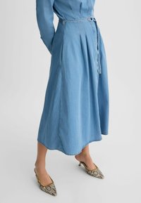 Hellblaues Jeanskleid mit tailliertem Schnitt, plissiertem Rock und kurzen Ärmeln. Kombiniert mit hochhackigen Schuhen im Schlangenlederdesign.
