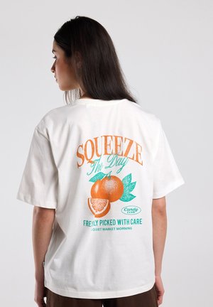 Woman with long dark hair seen from behind wearing a white T-shirt with orange and green fruit graphics and text "Squeeze The Day."