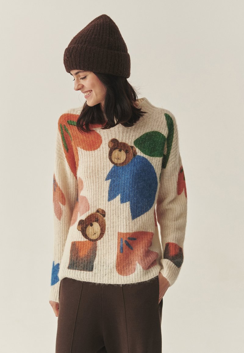 Pletena puli z barvito medvedjo in cvetličnimi vzorci v kremni barvi. Ima dolge rokave in sproščen kroj. V kombinaciji z rjavim rebrastim kapo.