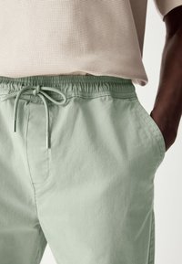 Pantalon vert clair à taille élastique avec deux poches latérales, cordon de serrage resserré et tissu en coton lisse. Chemise rayée visible au-dessus de la taille.