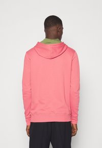 Homme de dos portant un sweat à capuche rose sur une chemise verte et un pantalon noir, sur un fond clair uni.