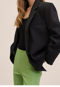 Vrouw in een zwarte blazer over een zwart top met een hoge taille in limoen groene broek, staand met de handen in de zakken tegen een neutrale achtergrond.