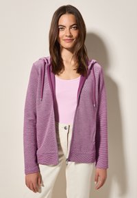 Veste à fermeture éclair violette en tissu texturé, avec capuche à cordon, poches avant et poignets côtelés. Portée sur un haut rose clair.