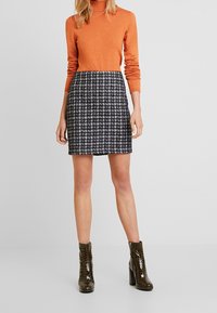 Pomarańczowy sweter z golfem w połączeniu z czarną i białą spódnicą w kratę, uzupełniony błyszczącymi, ciemnozielonymi botkami do kostek.