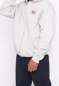Lichtgrijze zip-up hoodie, gemaakt van zachte stof, met een voorzak en geborduurd "MG" logo in kastanjebruin op de linker borst.