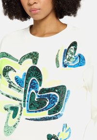 Biała bluza z dużymi cekinowymi wzorami serc w odcieniach zieleni, niebieskiego i żółtego. Gładka tekstura, okrągły dekolt i długie rękawy.