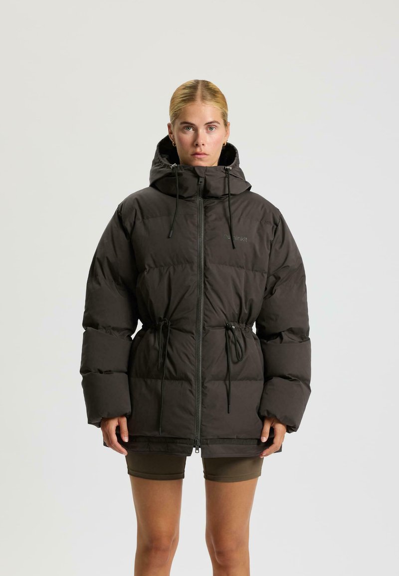 Eine Frau mit blonden Haaren trägt eine schwarze Steppjacke mit Kapuze, Kordelzügen und Reißverschluss, kombiniert mit olivgrünen Shorts, vor einem einfarbigen Hintergrund.