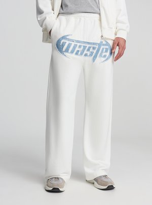 Pantaloni della tuta bianchi con logo ricamato blu, gambe larghe, vita elastica e texture morbida. Indossati con sneakers grigie.