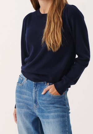 Kvinde iført en marineblå langærmet sweater med ribkantede ærmer og lyseblå højtaljede jeans, hånd i lommen.