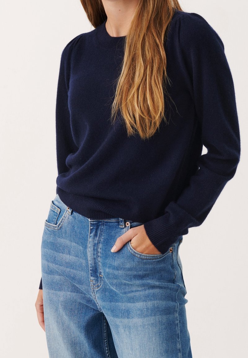 Femme portant un pull bleu marine à manches longues avec des poignets côtelés et un jean taille haute bleu clair, main dans la poche.