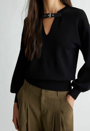 Mujer vestida con una blusa negra de manga larga con detalle de hebilla en el escote en V y pantalones plisados de talle alto en verde oliva, con la mano en el bolsillo.