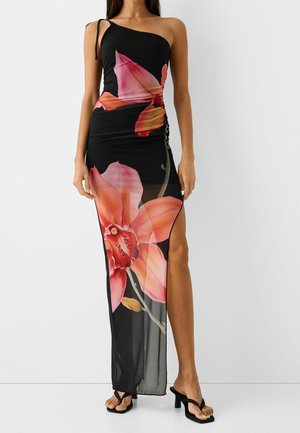 Femme portant une robe noire une épaule avec un grand imprimé floral rouge et rose et une fente haute, associée à des sandales noires à talons.