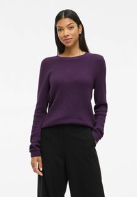Femme aux longs cheveux foncés portant un pull en tricot texturé violet et un pantalon noir, se tenant avec une main dans la poche sur fond blanc.