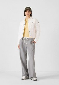 Giacca bianca trapuntata con chiusura a bottoni, camicia gialla, pantaloni larghi grigi e sneakers argentate; cappellino verde oliva con dettaglio del logo.
