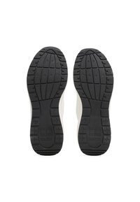 Semelles en caoutchouc noir de baskets blanches avec des motifs d'adhérence texturés et la marque "Tommy Jeans" au niveau du talon, vues par le dessous.