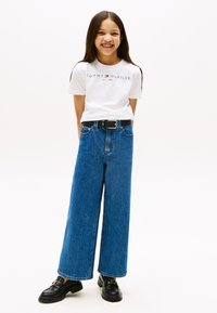 Bela bombažna majica s logotipom "Tommy Hilfiger EST. 1985", v kombinaciji z modrimi širokimi raztegnjenimi kavbojkami in črnimi loaferji z zlatim poudarkom.