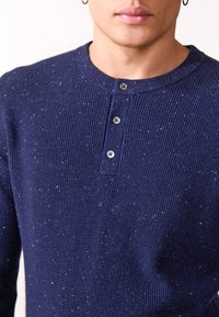 Tumedat sinise henley särk ribakujundusega, millel on kolm hõbedast nööpi ja ümar kaelus. Väikesed valged täpid lisavad detaili.