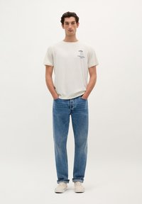 Hvid bomulds t-shirt med et trykt design på venstre bryst, parret med lyseblå lige ben jeans og beige sneakers.