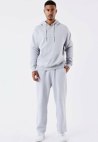 Sudadera gris claro con un bolsillo frontal y cordones, combinada con pantalones de chándal a juego. Ambos artículos están hechos de una tela suave; zapatillas blancas.
