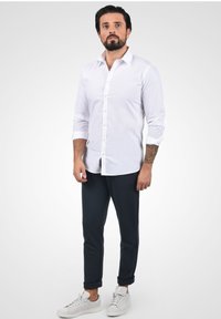 Hvit skjorte med lange ermer, kombinert med mørke bukser og hvite sneakers. Har en slim fit og oppbrettede ermekanter.