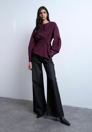 Donna con capelli lunghi e scuri che indossa una blusa bordeaux annodata, jeans neri a gamba larga e scarpe nere con la punta, in piedi contro un muro semplice.