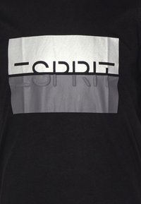 Černé tričko s stříbrným horizontálním designem a textem "ESPRIT" tučnými černými písmeny. Tkanina vypadá hladce a má mírně lesklou strukturu.