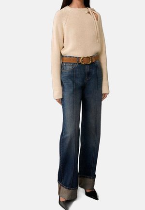 Femme portant un pull en maille crème avec un nœud sur l'épaule, un jean bleu foncé taille haute avec revers, une ceinture marron et des chaussures noires à bout pointu.