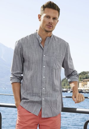 Grau gestreifte Hemd mit Knopfleiste und Stehkragen, einer einzelnen Brusttasche und langen Ärmeln, die an den Manschetten hochgerollt sind. Getragen mit korallenfarbenen Shorts.