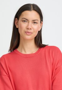 Pull de couleur corail avec un col rond et des manches longues. Le tissu semble doux avec une texture côtelée sur toute la surface.