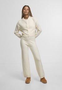 Krémově zbarvený pletený svetr na knoflíky a široké kalhoty ve stejném stylu. Jemná struktura; celkový vzhled doplňuje nenápadná obuv v neutrálních tónech.