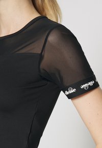 Černý sportovní top s průhlednými síťovanými rukávy. Má kulatý výstřih a elastické manžety s bílým logem. Úzký střih a jemná textura.