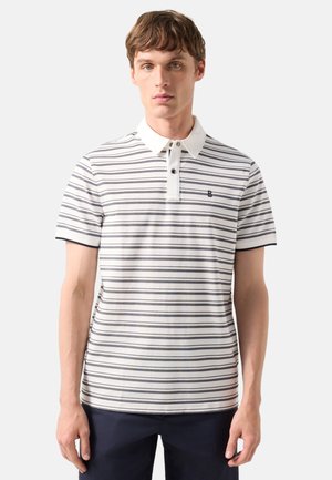 Junger Mann mit einem weiß und marineblau gestreiften Poloshirt mit weißem Kragen, steht vor einem schlichten hellen Hintergrund.