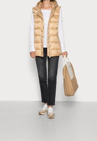 Beige pufferväst med huva, texturerad yta, över en vit långärmad skjorta. Mörka jeans och beige sneakers. Ljust brun tote-väska.