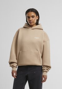 Beige hettegenser med en framre kenguru-lomme, ribbestrikkede mansjetter og en liten hvit logo på brystet. Laget av mykt, teksturert stoff.