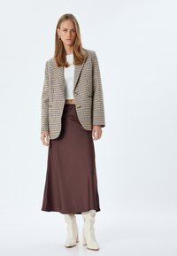 Blazer a motivo pied de poule in beige e marrone, top corto bianco, gonna lunga marrone e stivali crema al ginocchio, che mostrano un look a strati.