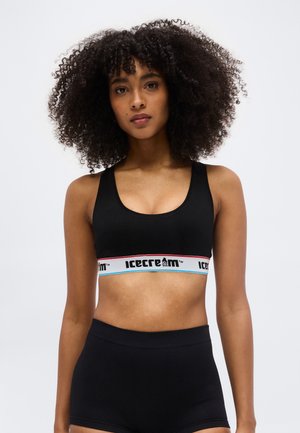 Jauna sieviete ar cirtainiem matiem, tērpusies melnā sporta krūšturī ar uzrakstu "icecream" un melnās augsta vidukļa šortos, skatās uz priekšu.