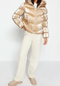 Giacca puffer color oro metallico con cappuccio, caratterizzata da un motivo a cuciture diagonali e tasche con zip. Abbinata a pantaloni larghi color crema e stivali beige.