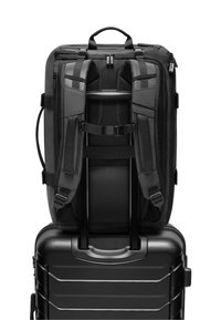 Mochila negra con correas ajustables, espalda de malla y asa superior, reposando sobre una maleta dura negra con superficie texturizada y asa moldeada.