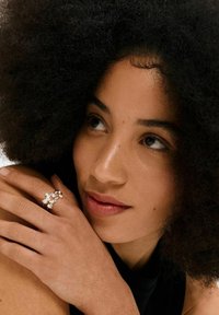 Silberring mit dekorativen Steinen, ausgestattet mit einem wellenförmigen Design. Modell hält den Arm mit strukturiertem Hautbild und lockigem, dunklem Haar. Dezentes Make-up.