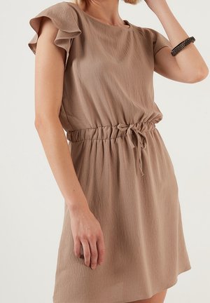 Femme portant une robe marron clair à manches courtes avec des manches volantées et une ceinture élastique nouée, accessoirisée d'un bracelet noir.
