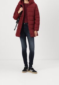 Rød polstret jakke med hætte, med et quiltet design og trykknapper. Kombineret med mørke jeans og sorte sko med hvide striber.