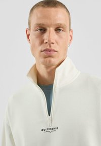 Biała bluza z suwakiem przy szyi, wykonana z miękkiego materiału, z małym czarnym logo oraz warstwową, jasnoniebieską koszulką pod spodem.